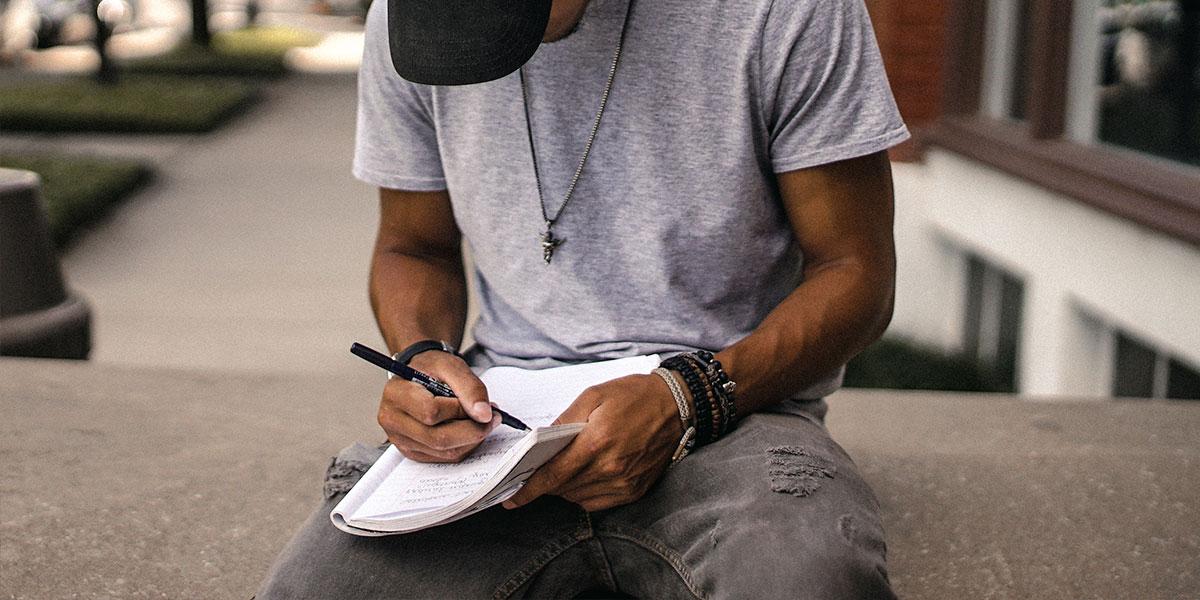 person in black adidas cap sitting on bench writing on notebook